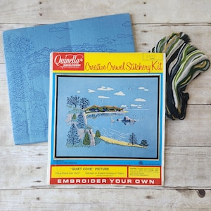 May include: A Quinella Needlework Creative Crewel Stitchery Kit titled Quiet Cove. The kit includes a blue fabric piece with a printed landscape outline, a bundle of green, white, and black yarn, and a box showing a finished coastal scene.