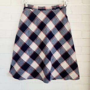 May include: A-line skirt with a plaid pattern in navy blue, white, and red. The skirt is hanging on a white hanger against a white brick wall. The plaid design features large squares and diagonal lines.