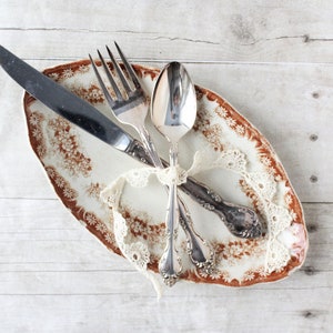 May include: An oval-shaped plate with a floral pattern in brown and white, accompanied by a knife, fork, and spoon. The silverware is tied together with a white lace ribbon. The arrangement is set on a light-colored wooden surface.