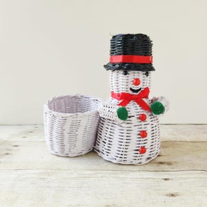 May include: A white woven snowman-shaped basket with a black top hat, red bow tie, and green mittens. A smaller, matching white basket sits beside it. The snowman has a red nose and red buttons. The baskets are on a light wood surface.