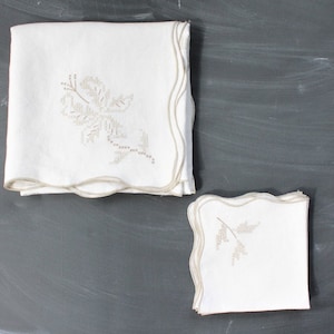May include: White linen napkins with a scalloped edge and embroidered floral design. The napkins are stacked, with one larger stack and a smaller stack of matching napkins. The embroidery is a light beige color.