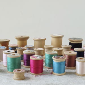 May include: A collection of vintage wooden spools wrapped in various colors of sewing thread, including shades of teal, magenta, blue, brown, and pink. The spools are arranged in a group, with some featuring paper labels on the ends.