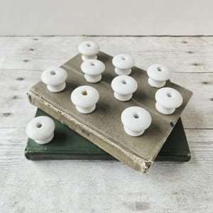 May include: A collection of ten small, white, mushroom-shaped ceramic knobs with a smooth, glossy finish. Each knob features a central hole and a rounded top, arranged in a cluster on top of two stacked, weathered, cloth-bound hardcover books.