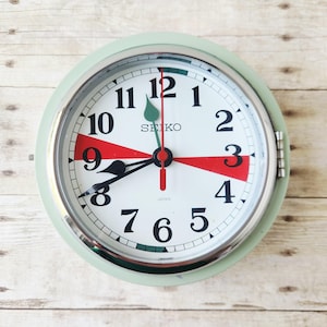May include: A round, mint green Seiko clock with a white face and black numerals. The clock features red accents at the 3 and 9 o'clock positions and a silver-colored bezel. The hands are black and red.
