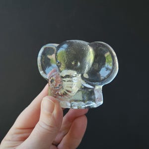 May include: A clear glass elephant figurine with large ears and a detailed trunk. The elephant is held in a hand against a dark background. The figurine is a decorative item.
