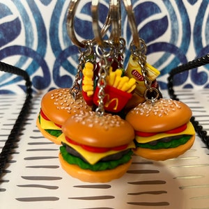 May include: A set of four miniature burger keychains with sesame seed buns, lettuce, tomato, cheese, and a meat patty. The keychains are attached to a silver chain with a bag of Lay's potato chips and a box of french fries.