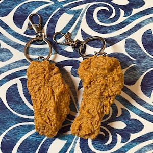 May include: Two fried chicken leg keychains with silver key rings. The keychains are made of a realistic-looking brown and yellow material.