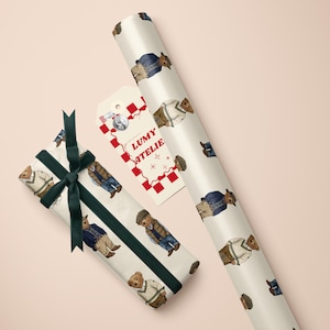 May include: Gift wrap featuring a repeating pattern of teddy bears in various outfits. A wrapped gift with a dark green ribbon and a tag with the text "LUMY ATELIE" are also visible. The background is a light pink.