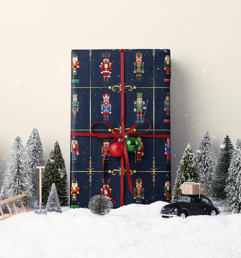May include: A wrapped gift with navy blue paper featuring a pattern of colorful nutcracker soldiers. The gift is tied with a red ribbon and adorned with a red and green ornament. Miniature Christmas trees and a toy car are in the foreground.