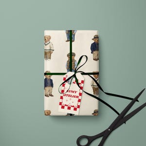 May include: Wrapped gift with a repeating pattern of teddy bears in various outfits. The gift is tied with a black ribbon and a tag that reads "LUMY ATELIER" in red lettering. A pair of black scissors is in the foreground.