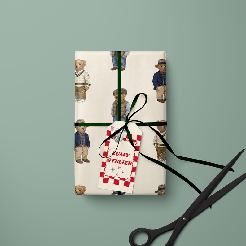 May include: Wrapped gift with a repeating pattern of teddy bears in various outfits. The gift is tied with a black ribbon and a tag that reads "LUMY ATELIER" in red lettering. A pair of black scissors is in the foreground.
