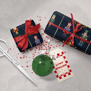 May include: Two wrapped gifts with red bows and nutcracker-themed paper, a green ornament, scissors, and a tag that says "LUMY ATELIER" with a red and white checkered border. Red star confetti is scattered around the items.