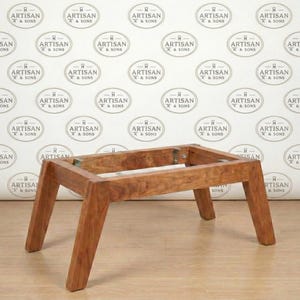 May include: A wooden table frame with four angled legs. The frame is made of light brown wood and has a rectangular top. The background features a repeating pattern of the words "ARTISAN & SONS" in a circular design.
