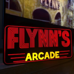 May include: A rectangular black sign featuring the words "FLYNN'S ARCADE" in red and yellow neon-style lettering. The sign has a retro design, with a dark background and bright, glowing text. It is likely for an arcade.