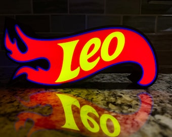 May include: A red, flame-shaped sign with the name "Leo" in yellow lettering. The sign has a blue outline and a black border. The sign is illuminated, and its reflection is visible on the surface below.
