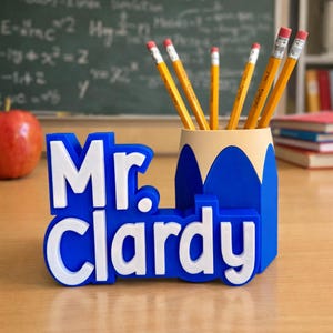 May include: A blue and white pencil holder with the name "Mr. Clardy" in bold letters. The pencil holder is shaped like a pencil and holds several yellow pencils with pink erasers. An apple and a chalkboard are in the background.