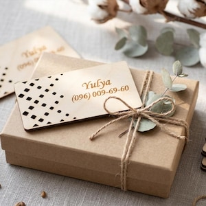 Puede incluir: Una caja de regalo de papel kraft atada con hilo, coronada con una tarjeta de visita de madera. La tarjeta presenta el nombre "Yulya" y un número de teléfono. Se ven tarjetas de madera adicionales con un patrón geométrico.