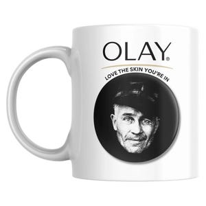 May include: White ceramic mug with a curved handle. The mug features the Olay logo in black, with the text "LOVE THE SKIN YOU'RE IN" below it. A black and white portrait of a man is on the mug.