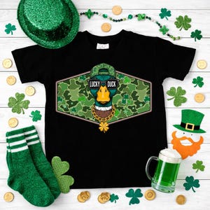 May include: Black t-shirt featuring a graphic of a duck wearing sunglasses, a camouflage hat, and a gold chain. The text "LUCKY DUCK" is above the duck. The shirt is surrounded by St. Patrick's Day decorations.