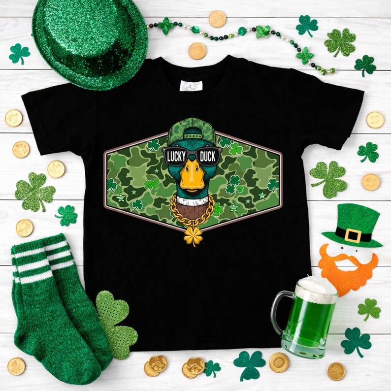 May include: Black t-shirt featuring a graphic of a duck wearing sunglasses, a camouflage hat, and a gold chain. The text "LUCKY DUCK" is above the duck. The shirt is surrounded by St. Patrick's Day decorations.