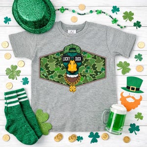 May include: A gray t-shirt featuring a graphic of a duck wearing sunglasses, a hat, and a gold chain, with the text "LUCKY DUCK". The shirt is surrounded by St. Patrick's Day decorations, including shamrocks, gold coins, and green socks.