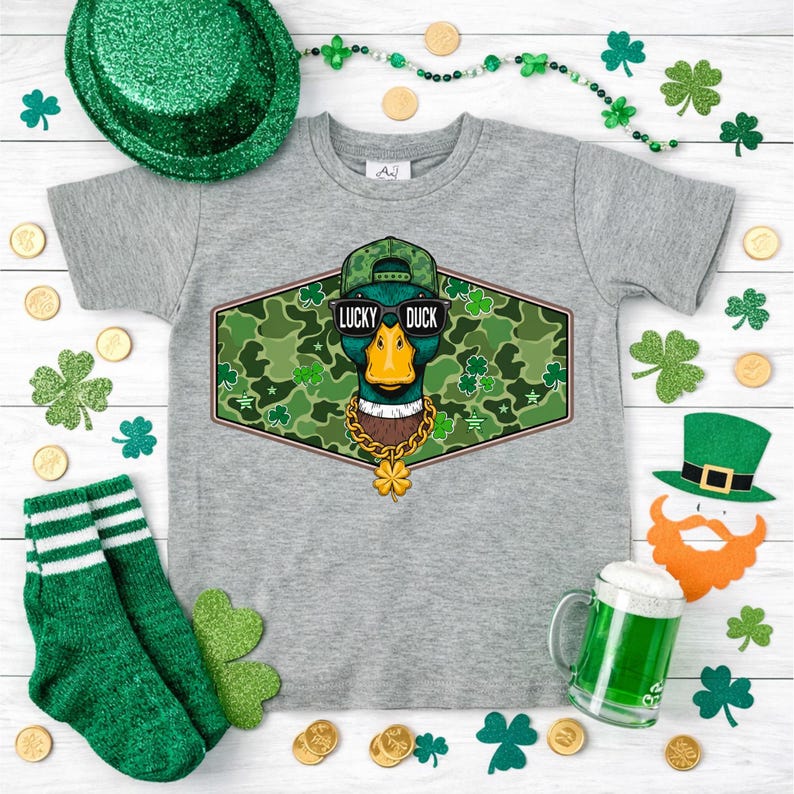 May include: A gray t-shirt featuring a graphic of a duck wearing sunglasses, a hat, and a gold chain, with the text "LUCKY DUCK". The shirt is surrounded by St. Patrick's Day decorations, including shamrocks, gold coins, and green socks.