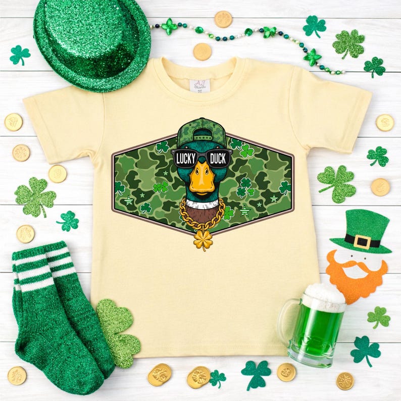 May include: A light yellow t-shirt featuring a graphic of a duck wearing sunglasses, a hat, and a gold chain. The graphic is surrounded by a camouflage pattern and the words "Lucky Duck". Green socks, shamrocks, and gold coins are scattered around the shirt.