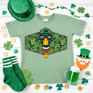 May include: A light green t-shirt with a graphic of a duck wearing sunglasses, a hat, and a gold chain. The text "LUCKY DUCK" is above the duck. The shirt is surrounded by St. Patrick's Day decorations, including shamrocks and a green hat.