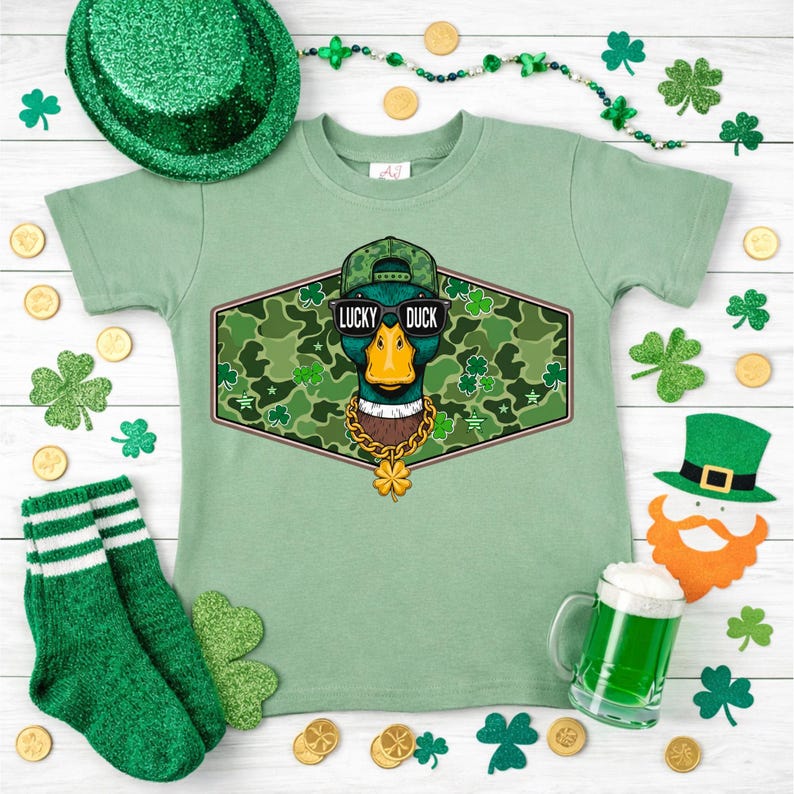 May include: A light green t-shirt with a graphic of a duck wearing sunglasses, a hat, and a gold chain. The text "LUCKY DUCK" is above the duck. The shirt is surrounded by St. Patrick's Day decorations, including shamrocks and a green hat.