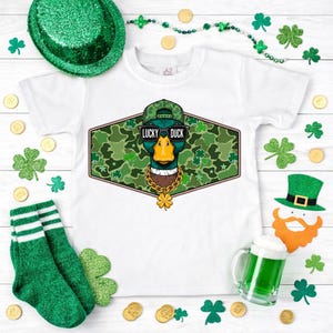 May include: White t-shirt featuring a cartoon duck wearing sunglasses, a cap, and a gold chain, with the text "LUCKY DUCK". The design is set against a green camouflage background, with shamrocks and St. Patrick's Day decorations.