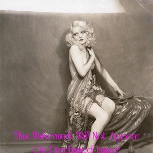 May include: A black and white vintage photograph of a woman with blonde hair wearing a sheer, patterned dress and a long, flowing scarf. She is sitting on a stool with her legs crossed and looking off to the side.