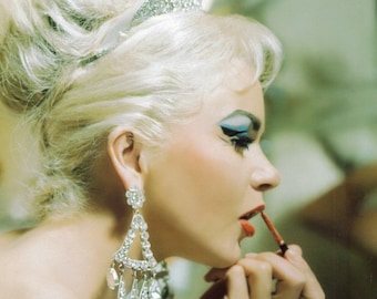 May include: A woman with blonde hair styled in a high bouffant wears a tiara and large diamond earrings. She is applying red lipstick with a brush.