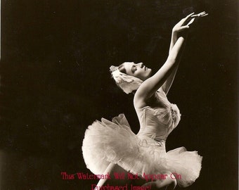 Op de afbeelding: Een zwart-witfoto van een ballerina die een witte tutu en een tiara draagt, poserend met haar armen boven haar hoofd.
