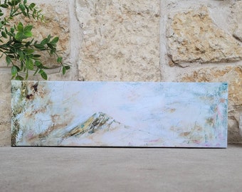 May include: Abstract landscape painting on a rectangular canvas. The artwork features a mountain range with a textured, layered effect. Colors include white, blue, gold, and touches of pink and green. The painting is displayed on a stone surface.