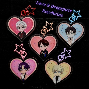 Op de afbeelding: Collectie Love & Deepspace sleutelhangers. Elke sleutelhanger toont een stripfiguur in een hartvormig ontwerp dat lijkt op een donut met hagelslag. De sleutelhangers hebben stervormige sluitingen in verschillende kleuren.