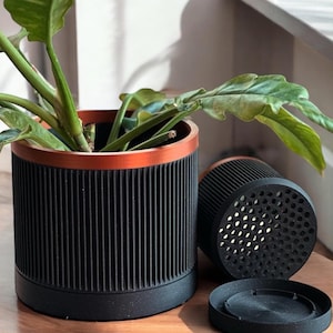 May include: A black and copper-coloured cylindrical planter with vertical grooves, holding a green plant. A second planter and a detached base are next to it. The planter has a copper-coloured rim.