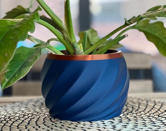 May include: A blue planter with a copper rim, featuring a swirl design, holds a green plant with large leaves. The planter is sitting on a woven straw placemat.
