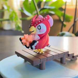 May include: A red owl figurine with white eyes and a pink beak holding a yellow star. The owl is sitting on a brown wooden dock with a blue water base.