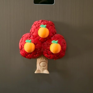 May include: A whimsical, three-dimensional tree-shaped magnet. The tree features a vibrant red canopy with three orange fruits, each with a green stem. The trunk is beige, and the magnet is set against a neutral background.
