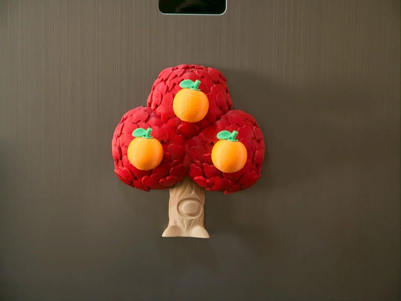 May include: A whimsical, three-dimensional tree-shaped magnet. The tree features a vibrant red canopy with three orange fruits, each with a green stem. The trunk is beige, and the magnet is set against a neutral background.