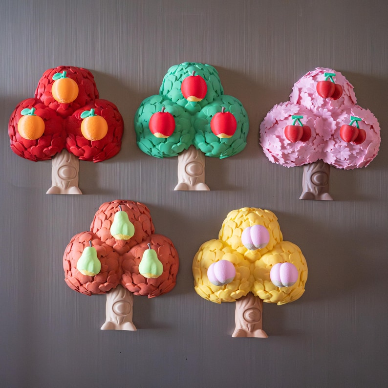 May include: A collection of six colorful tree-shaped magnets. The trees feature red, green, and pink foliage with orange, red, and cherry-like fruit. The trunks are a light beige color, and the magnets are arranged on a gray surface.