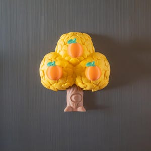 May include: A yellow tree-shaped magnet with three orange fruit. The fruit have green stems. The tree trunk is light brown. The magnet is on a gray background.