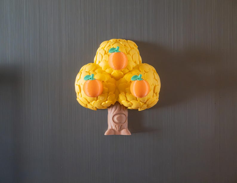 May include: A yellow tree-shaped magnet with three orange fruit. The fruit have green stems. The tree trunk is light brown. The magnet is on a gray background.