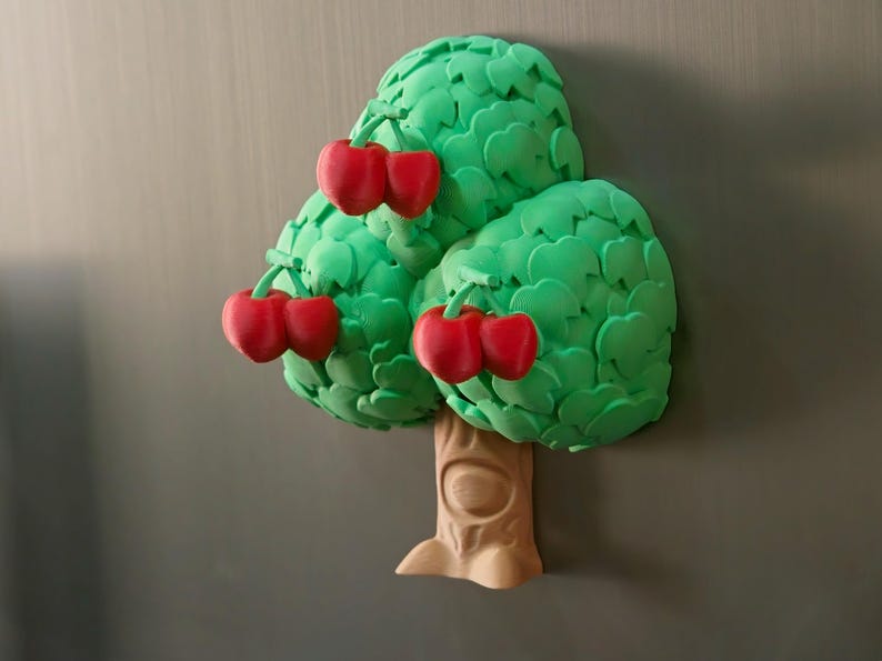 May include: A whimsical, three-dimensional tree magnet with a light brown trunk and vibrant green leaves. Clusters of bright red cherries adorn the tree. The magnet is designed to resemble a cartoon-like illustration.