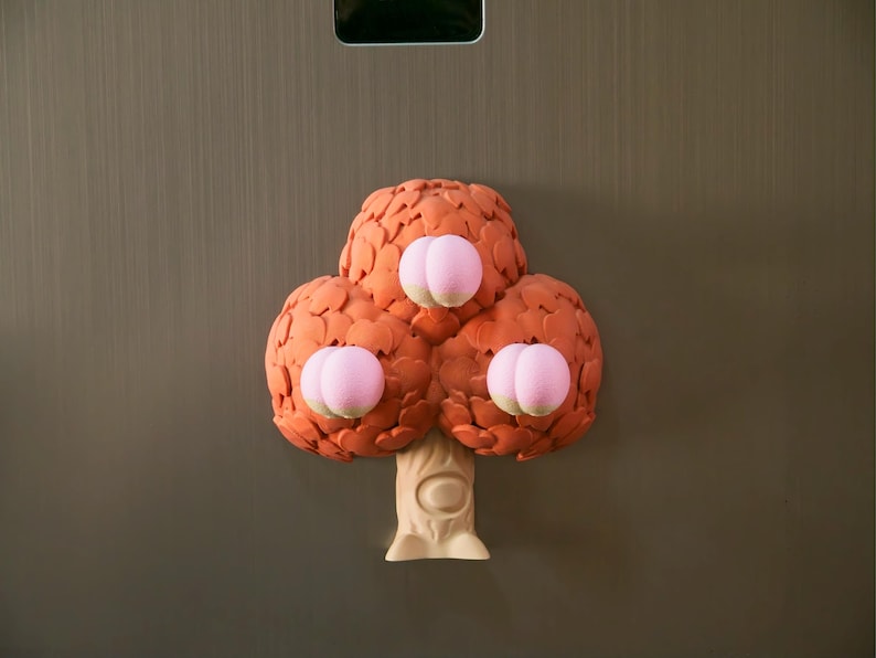 May include: A whimsical, three-dimensional tree sculpture with a light beige trunk and three orange-brown, spherical clusters of leaves. Each cluster features a pair of pink, rounded accents, creating a playful, cartoonish aesthetic. The sculpture is mounted on a gray surface.