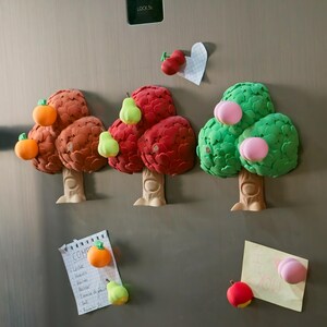 May include: A collection of colorful, fruit-themed refrigerator magnets. The magnets are shaped like trees with various fruits, including oranges, pears, and peaches. A notepad with handwritten text and a small heart-shaped note are also attached.