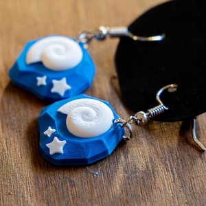 Puede incluir: Un par de pendientes azules y blancos. Cada pendiente presenta una base azul facetada con un diseño en espiral blanco y dos estrellas blancas. Los pendientes tienen ganchos plateados y están sobre una superficie de madera.