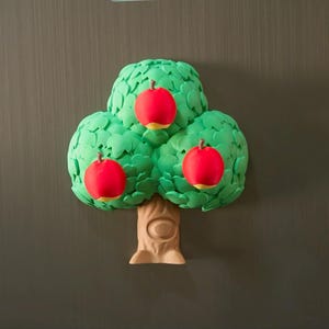 May include: A whimsical tree-shaped magnet with three red apples. The tree has a green, leafy canopy and a tan trunk. The apples have yellow bottoms. The magnet is likely for a refrigerator or other metal surface.