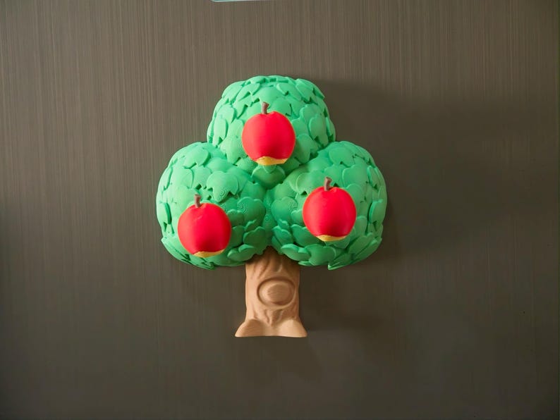 May include: A whimsical tree-shaped magnet with three red apples. The tree has a green, leafy canopy and a tan trunk. The apples have yellow bottoms. The magnet is likely for a refrigerator or other metal surface.