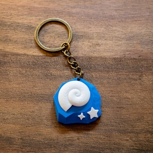 Può includere: Un portachiavi blu con un design a conchiglia bianca e due stelle bianche. Il portachiavi ha un anello e una catena color bronzo. Lo sfondo è una superficie di legno.